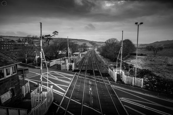 Monochrome shot over railway lines leading into the distance. Signal box and crossing in shot, heavy sky above