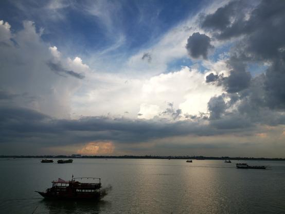 Photo shows a late afternoon photo of the meeting point of two large river water bodies in the lower half of the photo. Some boats in different sizes are spread out going in different directions.  In the middle is a strip of land with some buildings, including some high rises. In the upper half of the photo there is a changing sky mix of deep, clear blue, light white clouds of a light, hot day, and deep dark and and rain heavy cloud in the distance with a glimpse of yellow clear light being the sun nearing its setting phase.