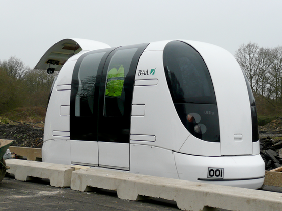 An Ultra PRT vehicle on a test track at Heathrow Airport, London. It's a very small white rounded train car that opens at the back for luggage and has two double doors on the side to let just a handful of people in. You can see someone in a high vis safety vest inside tinted doubledoor windows.