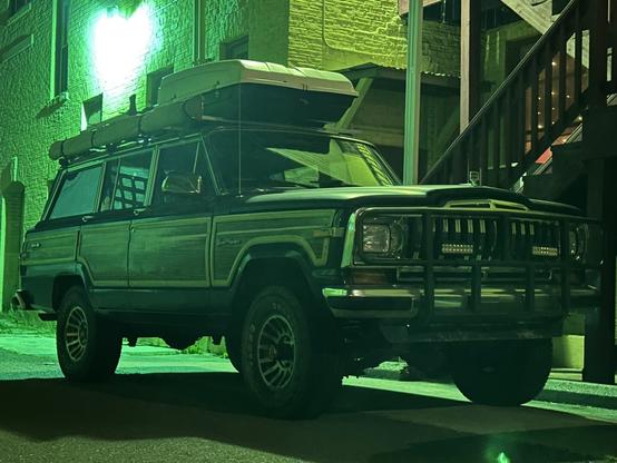 A vintage SUV parked in an alleyway, illuminated by green lighting. The vehicle has a rack holding gear on the roof, with a classic wood panel design and rugged tires. The surrounding area features brick walls and a staircase, contributing to an urban atmosphere
