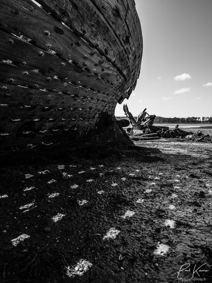 Photo monochrome en noir et blanc en format vertical, présentant des parties d'épaves de bateaux dans un cimetière dédié à leur fin de vie en haut d'une grève, à marée basse. Sur les deux tiers gauche de la photo, on peut voir le flanc d'une carène (et son ombre) où le calfatage entre les bordés a depuis longtemps disparu, laissant ainsi filtrer la lumière entre les membrures. 
Plus loin, à droite, on perçoit les derniers restes de la quille et du moteur d'un autre bateau, la décrépitude de celui-ci étant nettement plus avancée. Dans le ciel dans le coin supérieur droit, seuls trois tout petits nuages sont visibles.