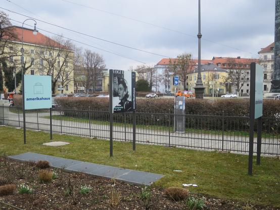 Das Bild zeigt eine öffentliche Parkanlage mit mehreren Schildern und einem Metallzaun. Im Vordergrund befindet sich ein Schild mit der Aufschrift "amerikahaus" und einem Gebäude-Logo. Neben ihm steht ein großes Schild mit einem Schwarz-Weiß-Bild einer Frau und der Aufschrift "VOGUE". Im Hintergrund sind Gebäude mit roten Dächern und ein gepflegter Rasen zu sehen. Einige Autos fahren auf der Straße dahinter. Der Himmel ist bewölkt, und es gibt ein paar Bäume ohne Blätter, was auf eine kühle Jahreszeit hinweist.