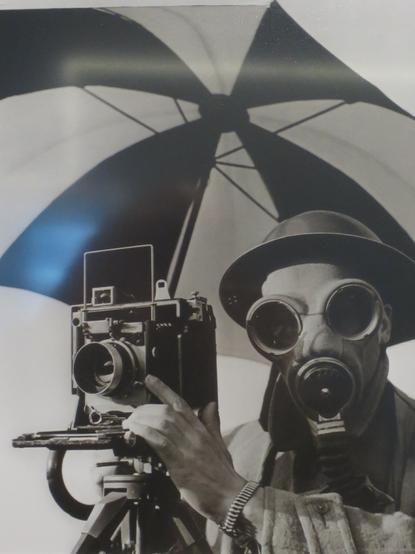Das Bild zeigt einen Mann, der eine alte, große Kamera auf einem Stativ bedient. Er trägt eine Gasmaske und eine breite, dunkle Mütze. Die Kamera ist ein klassisches Modell mit einem großen Objektiv und einem Reflexkasten. Der Mann hat eine besondere Konzentration auf das Objektiv gerichtet, während er die Kamera einstellt. Im Hintergrund ist ein großes, schwarz-weißes Regenschirm aufgeschlagen, das Licht reflektiert und einen starken Kontrast erzeugt. Die gesamte Szene ist in Schwarz-Weiß gehalten, was eine zeitlose Atmosphäre vermittelt.