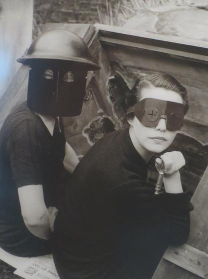 Das Bild zeigt zwei Personen in einem schwarz-weißen Format. Die Person auf der linken Seite trägt einen dunklen, metallischen Helm mit zwei runden Augenlöchern, der an einen Soldatenhelm erinnert. Sie sitzt mit dem Rücken zur Kamera und trägt ein dunkles, kurzärmeliges Oberteil. Die Person auf der rechten Seite trägt eine Art Visier mit Kreuzen an den Augenöffnungen und hält einen Gegenstand in der Hand, der wie ein Schlüssel aussieht. Sie sitzt mit dem Oberkörper leicht nach vorne geneigt und trägt ein dunkles, langärmeliges Oberteil. Beide Personen sitzen in einem schmutzigen, hölzernen Behälter, der an eine Schanze oder ein Versteck erinnert. Im Hintergrund ist ein bröckelnder Steinmauerabschnitt zu sehen. Die Gesamtkomposition des Bildes vermittelt eine düstere und mysteriöse Atmosphäre.