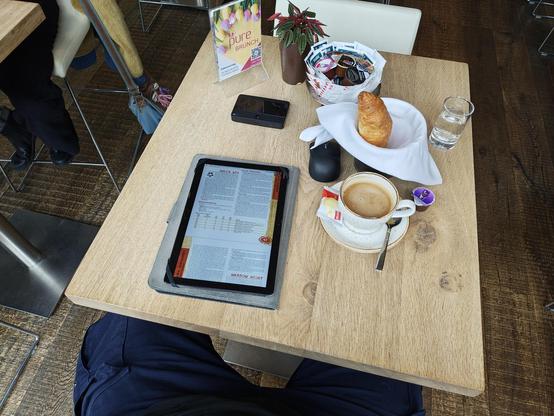 Tablet mit PDF, Tasse Kaffe, Croissant auf einem hellen Tisch aus Holz