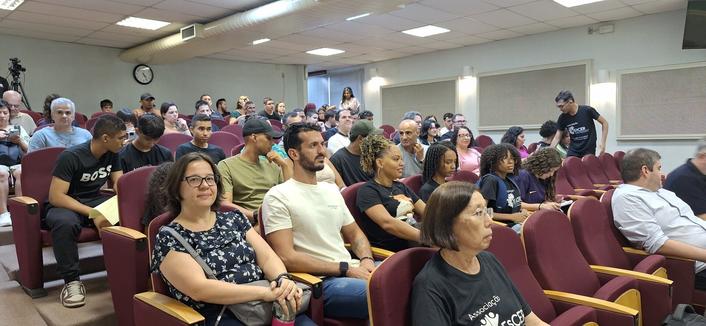 A imagem mostra um auditório com várias fileiras de assentos vermelhos, ocupados por um grupo diverso de pessoas. A maioria está sentada, com alguns em pé, todos voltados para a frente, indicando que estão assistindo a uma apresentação ou palestra. As pessoas estão vestidas de maneira casual. A sala é iluminada por luzes fluorescentes no teto, e há uma grande janela com cortinas brancas no fundo. Um relógio está visível na parede, mostrando a hora. A atmosfera parece ser de concentração e interesse, com a maioria das pessoas olhando para a frente.