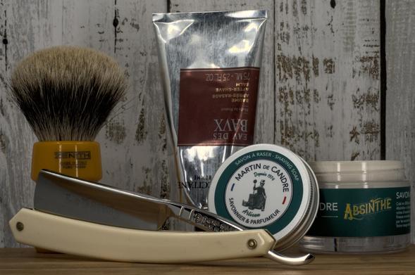 (Le Chat Mistral description)

This image features a collection of men's grooming items arranged on a rustic wooden surface. In the foreground, there's a straight razor with a white handle and a silver blade, engraved with "Le Dandy." To the left, a shaving brush with a yellow and black handle and light bristles stands upright. Behind it, a metallic tube of "Eau des Baux" shaving balm, 75 millilitres, is visible. To the right, a round tin of "Martin de Candre" shaving soap in the "Absinthe" scent is displayed, evoking a classic barbershop aesthetic.