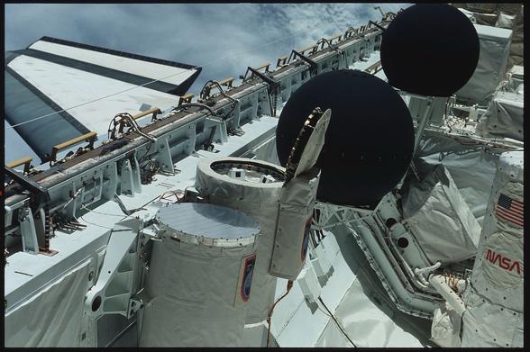 The image features the exterior of a space shuttle with various instruments and components visible. Notable elements include two large black spherical objects and several cylindrical devices covered in protective fabric. The NASA logo and an American flag are also present on the shuttle's surface.