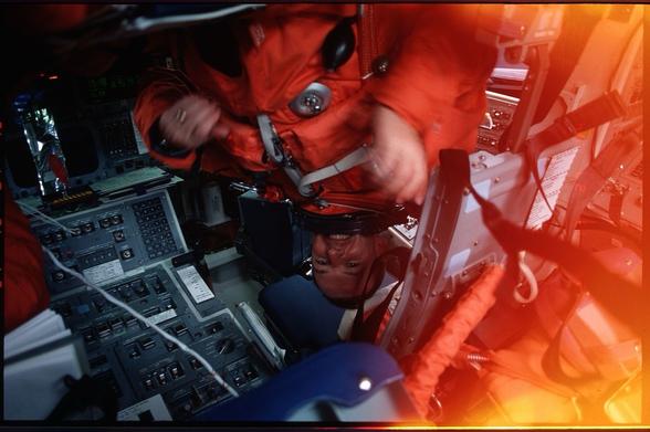 An astronaut in an orange spacesuit is inside a spacecraft, smiling and operating controls. The cockpit displays various instruments and buttons, with some wires and equipment visible. The image has a warm, orange tint.