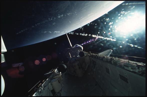 A view from space showing the curved horizon of Earth illuminated by sunlight, with a spacecraft in the foreground. Stars and light reflections create a celestial atmosphere.