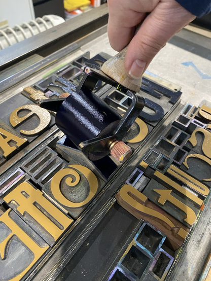 Photo of a letterpress lockup showing a hand with a tollercapplying ink to the wood type letters