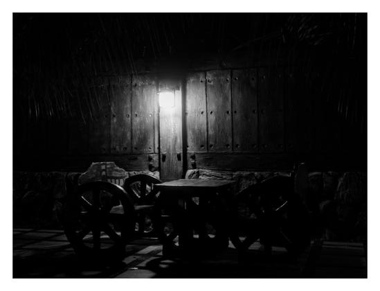 A black and white photograph of an outdoor setting at night. There's a wooden gate or wall illuminated by a wall lantern. In front of it, there is a bench with large, wagon-wheel-like elements on the sides, all silhouetted against the light. The scene is framed by what appears to be wooden walls on either side. - Google Gemini