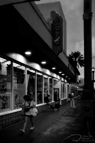 A street with a shop front and neon sign of a liquor and tobacco shop at dusk. A few people are hanging around the entrance, a young woman with a bag walks out of the picture on the left.