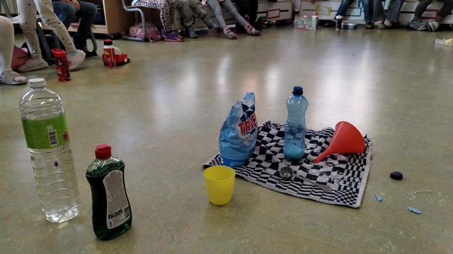 Een kring van kindervoeten en op de grond staat een 0,5L flesje, een fles afwasmiddel, een fles azijn, een zak zilversoda en een trechter.