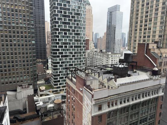 Urban skyline view featuring a mix of modern and older buildings, including high-rise structures, rooftop water tanks, and green spaces, under cloudy skies.