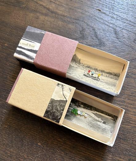 Two matchboxes are displayed side by side. One box is covered with a collage of brown paper and a black-and-white photograph of a lake. Inside, it features a black-and-white photograph of a small boat on the water with three people on board, their faces covered with colorful beads. The other box is covered with a collage of beige paper and a black-and-white photograph of a tree and a mountain. Inside, it displays a black-and-white landscape photograph accented with several beads.