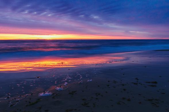 A breathtaking sunset over a Danish beach, where vivid shades of orange, pink, and purple reflect on the wet sand and waves roll gently onto the shore.