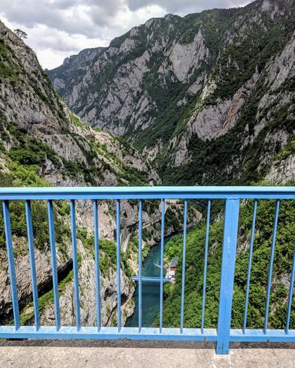 A wide, slightly high-angle shot shows a dramatic mountain gorge with a turquoise river flowing through it, as seen from a bridge. The foreground features a bright blue metal railing of the bridge, which runs horizontally across the lower portion of the frame. The bridge itself has a grey concrete surface visible just below the railing.
Beyond the railing, the view plunges down into a deep canyon. Steep, rocky mountain slopes rise on either side, covered in patches of green vegetation, mostly trees and shrubs. The rock faces are a mix of grey and light brown, showing signs of erosion and geological layering.
At the bottom of the gorge, a vibrant turquoise river snakes its way through. A small, white structure with a dark roof appears to be situated on the riverbank or perhaps a small island within the river. The water reflects the cloudy sky above, which is visible in patches between the mountain peaks. The overall lighting suggests an overcast day.