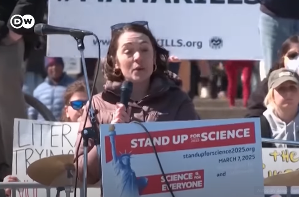 Frau spricht am Mikrofon bei einer Demonstration für Wissenschaft, mit einem Schild "STAND UP FOR SCIENCE" im Vordergrund. Im Hintergrund sind Teilnehmer mit Schildern zu sehen.