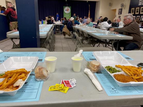 Color image of two boxed meals on a table in a church cafeteria. Image shows two opened meal boxes filled with food from a church social hall Fish Fry in Akron, Summit County, Ohio, USA, with long tables for diners. These are common in the USA during the Easter Lenten season on Fridays, called "Friday Fish Frys", with churches and charitable organizations using them as fundraisers during the Easter season.