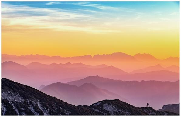 Silhouetted mountain peaks in the foreground give way to multiple layers of progressively lighter, misty mountains beneath a vibrant sunset or sunrise sky. A tiny figure of a person stands on a distant peak.