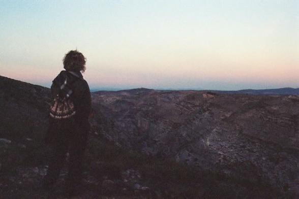 Una mujer de espaldas, mira al horizonte. Lleva el pelo corto con melena hasta el cuello y una mochila blanca y negra. Al fondo una montañas de piedra y un atardecer con todnos violetas y azules.
