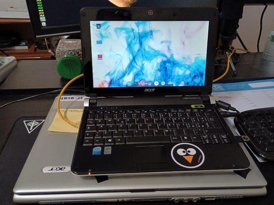 Piccolo netbook Acer acceso, appoggiato sopra un altro PC piu datato, si vede il piano di una scrivania con vari oggetti che potremmo trovare in un studio o laboratorio