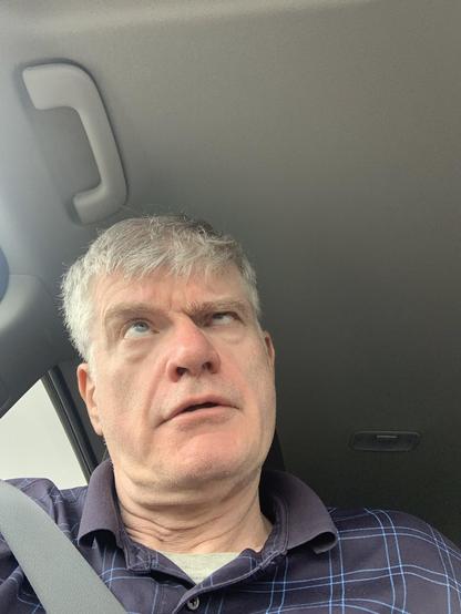 Photo of Martin Nutty with short gray hair, light skin, and a playful expression is sitting in a car, wearing a navy blue plaid polo shirt. He is rolling his eyes upward in an exaggerated manner, creating a comical, exasperated look. The seatbelt is fastened across his chest, and the car’s interior, including a handle on the ceiling, is visible in the background.
