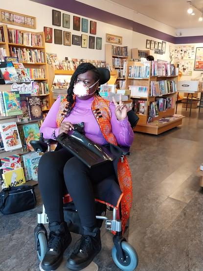 Jay in her wheelchair at Comix Experience, a comic book store with wooden shelves full of books and art on the walls. She is wearing a long bright orange vest over a purple shirt and a matching Flo Mask.