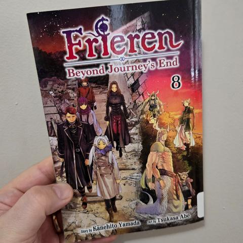 Photograph of a white person's hand holding Frieren: Beyond Journey's End, Volume 8 against a light colored wall. The title is at the top. The cover shows a group of three fantasy adventurers walking toward the viewer downhill with a long, rocky countryside behind them at night. On the right side is a monster with four arms and a snake lower half and two demons alongside them at dusk. The titular Frieren (white haired, long eared, short elf) is at the center standing toward the front of the group walking downward.