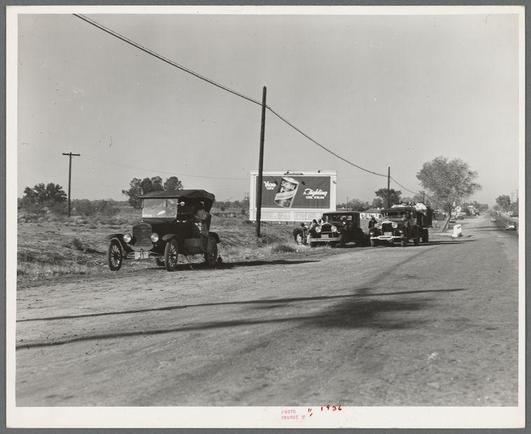 A black and white photograph depicting a rural roadside scene with three vintage automobiles parked on the side of an unpaved road. The vehicle closest to the foreground appears to be a Model T, identifiable by its boxy shape and prominent front grille. Behind it is another early automobile model, followed further back by yet another car from roughly similar era. On the right, there's also what seems like part of a person standing beside one of these vehicles.

A large billboard with an advertisement for a coffee brand looms in the background under clear skies. The road stretches into the distance with no visible traffic or pedestrians aside from those associated with the parked cars and roadside figures. There are utility poles along the side, suggesting some level of infrastructure development beyond what is immediately apparent.