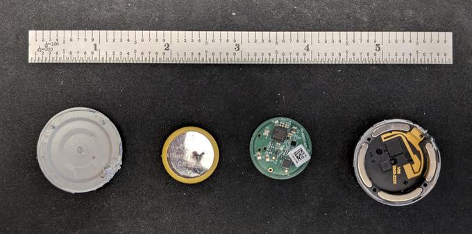 The covers, battery, and circuit board for a Princess Cruises tracking medallion on a matte black background with a steel 6" ruler for scale