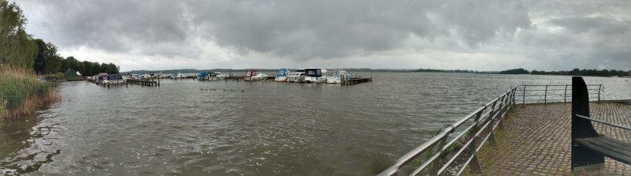 A lake under a grey sky with low hanging clouds. The panorama has a shore to its left, a jetty with many small boats, and this view from a terrace that is extending over the water. General atmosphere is that of a quiet moment in unstable summer weather.