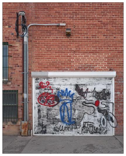 This image features a white garage door with graffiti art painted on it, set against a brick building. The graffiti includes a depiction of what appears to be a whimsical moose character in gray with a red nose, a crown-like symbol in blue, and an abstract red design resembling a brain with eyes. Adjacent to the garage door, there's an exposed pipe and electrical setup on the brick facade, adding an industrial feel to the scene. The brick wall also shows various signs of aging and wear, contributing to a gritty, urban aesthetic.
