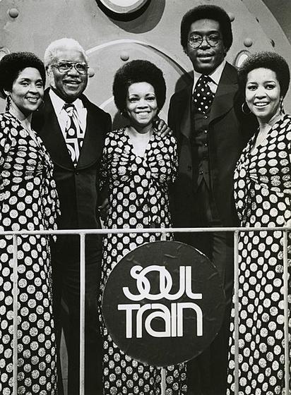 The Staple Singers in a black and white picture as they appeared on Soul Train on June 8, 1974. They're standing in a row. Pops is second from the left between his daughters Cleotha and Yvonne. TV host Don Cornelius is second from the right. Mavis is next to him.