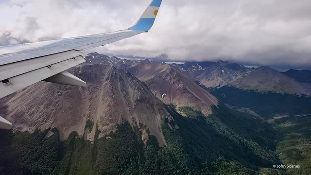 A wing over grey mountain peaks with patches of snow that turn to green foliage.  There's a glimpse of the Beagle Channel with grey skies above.