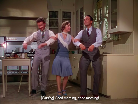 The three people are now dancing in the kitchen. The woman wears a dress with a swingy skirt, and her arms are linked with a white man on either side. Both of them are wearing suit vests and trousers. They're all smiling as they lift their feet to begin the dance. “[Singing] Good morning, good morning!”
