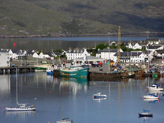 Ullapool in Wester Ross. The image shows a telephoto view of the harbour area of Ullapool, with moored fishing boats. There are smaller boats moored to buoys in the foreground, while in the background the white buildings of Ullapool give way to a loch, on the far side of which the land climbs out of the frame.