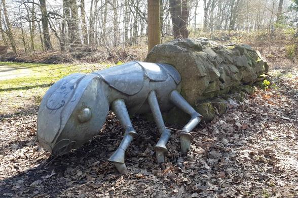 Ein Insekt, ähnlich einer Ameise, aus Metall ist mit Kopf und sechs Beinen zu sehen. Der Unterleib steckt in einem steinigen Köcher. Die einzelnen Steine der Behausung sind etwas abstehend zu erkennen.