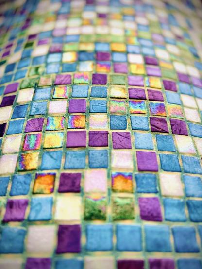 Mosaic tiles of white and colors violet, sky blue, emerald green, and patina gold arrange in a grid pattern up the side of a large outdoor column.