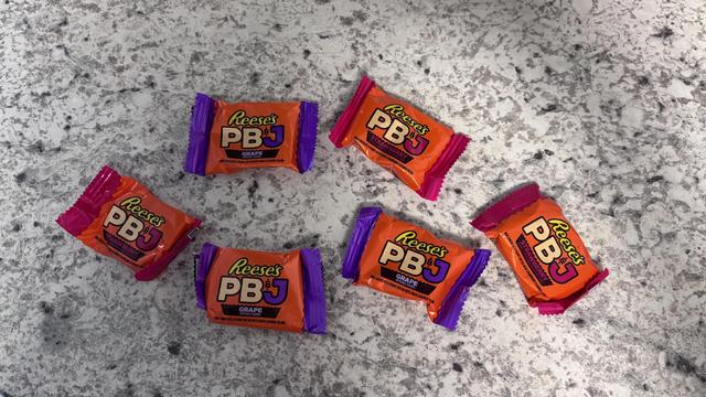 The image shows seven individually wrapped Reese's PB&J candies arranged on a speckled gray and white countertop. The wrappers are predominantly orange with purple and pink accents. Each wrapper features the text "Reese's PB&J" in white and yellow letters, with "GRAPE" written in smaller text below. The candies are positioned in a somewhat scattered pattern, with some facing different directions. The background is a light-colored, speckled surface, providing a neutral backdrop that contrasts with the vibrant colors of the candy wrappers.
