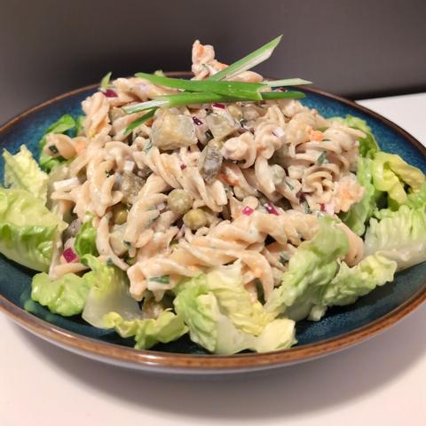 Photo of a pasta salad made of wholegrain fusilli pasta, pickled gherkins, red onion, green peas, carrot and chives with a vegan mayonnaise dressing. Around the salad there are light green roughly chopped lettuce leaves. The salad is garnished with four thin slices of spring onion and presented on a blue plate. The background is a neutral white table.