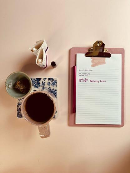 Eine Draufsicht auf eine rosa Tasse mit Tee, die auf einem blau-weißen Untersetzer steht, einer kleinen Schale mit einem Teebeutel, zwei Tintentuben, einer kleinen Flasche und einem Klemmbrett mit liniertem Papier und dem Lumos Pro Duo. Auf dem Papier steht, dass zum Schreiben die 0.3 writing tip mit Tinte in der Farbe Iris und die Brush Tip mit der Farbe Raspberry Sorbet benutzt wurden.