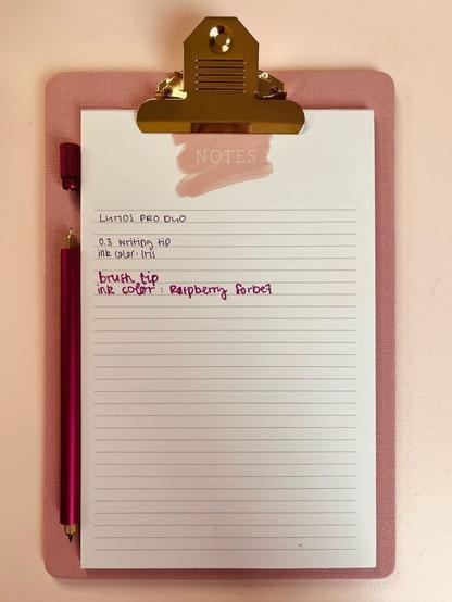 Ein rosa Klemmbrett mit einem linierten Notizblock zeigt handschriftliche Notizen mit Angaben zu einer 0,3 writing tip in der Tintenfarbe "Iris" und der brush tip in "Raspberry Sorbet". Daneben liegt der Lumos Pro Duo mit den beschriebenen Tips.