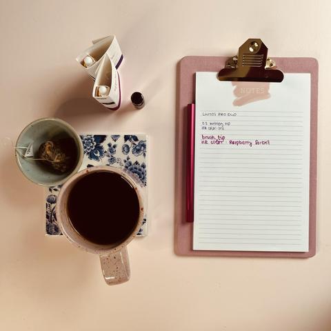 Blick von oben auf eine rosa Tasse mit Tee, die auf einem blau-weißen Untersetzer steht, einer kleinen Schale mit einem Teebeutel, zwei Tintentuben, einer kleinen Flasche und einem Klemmbrett mit liniertem Papier und dem Lumos Pro Duo. Auf dem Papier steht, dass zum Schreiben die 0.3 writing tip mit Tinte in der Farbe Iris und die Brush Tip mit der Farbe Raspberry Sorbet benutzt wurden.