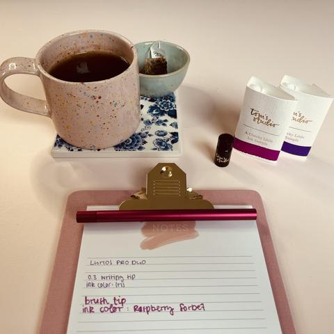 Blick von schräg oben auf eine rosa Tasse mit Tee, die auf einem blau-weißen Untersetzer steht, einer kleinen Schale mit einem Teebeutel, zwei Tintentuben, einer kleinen Flasche und einem Klemmbrett mit liniertem Papier und dem Lumos Pro Duo Stift. Auf dem Papier steht, dass zum Schreiben die 0.3 writing tip mit Tinte in der Farbe Iris und die Brush Tip mit der Farbe Raspberry Sorbet benutzt wurden.