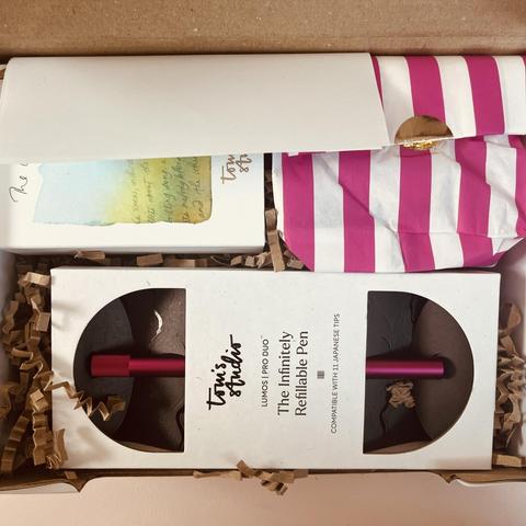 Blick von oben auf ein geöffnetes Paket. Man sieht eine Schachtel mit dem “Infinitely Refillable Pen” Lumos Pro Duo von Tom’s Studio in pink, eine kleinere Schachtel, die halb verdeckt ist und eine rosa-weiß gestreifte Papiertüte, die mit einem runden goldenen Sticker verschlossen ist.