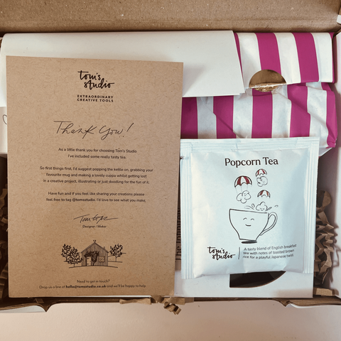 Blick von oben auf ein geöffnetes Paket. Obenauf liegen ein Beutel “Popcorn Tea” und eine Karte von Tom’s Studio, auf der die Aufforderung steht, sich erstmal in Ruhe eine Tasse Tee zu kochen und dann in einem kreativen Projekt zu versinken.
