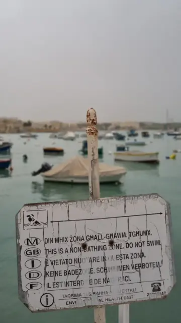 Schild "Keine Badezone. Schwimmen verboten!" am Hafen von Birżebbuġa. Im Hintergrund Wasser und dutzende Boote darauf