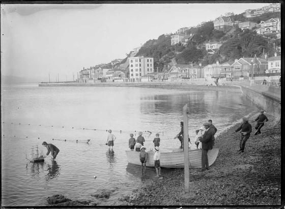 Black-and-white photo: Photograph of a scene at Oriental Bay, Wellington, including a boy with a model ship, and fishermen. 1922–ca. 1929. Photographer unknown. Description: A scene at the shoreline of Oriental Bay, Wellington, showing a boy with a model ship and fishermen holding a net. The background shows the hillside with buildings at the right and the harbour to the left. Citation: Ref: 1/2-075160-G. Alexander Turnbull Library, Wellington, New Zealand. https://natlib.govt.nz/records/22674925